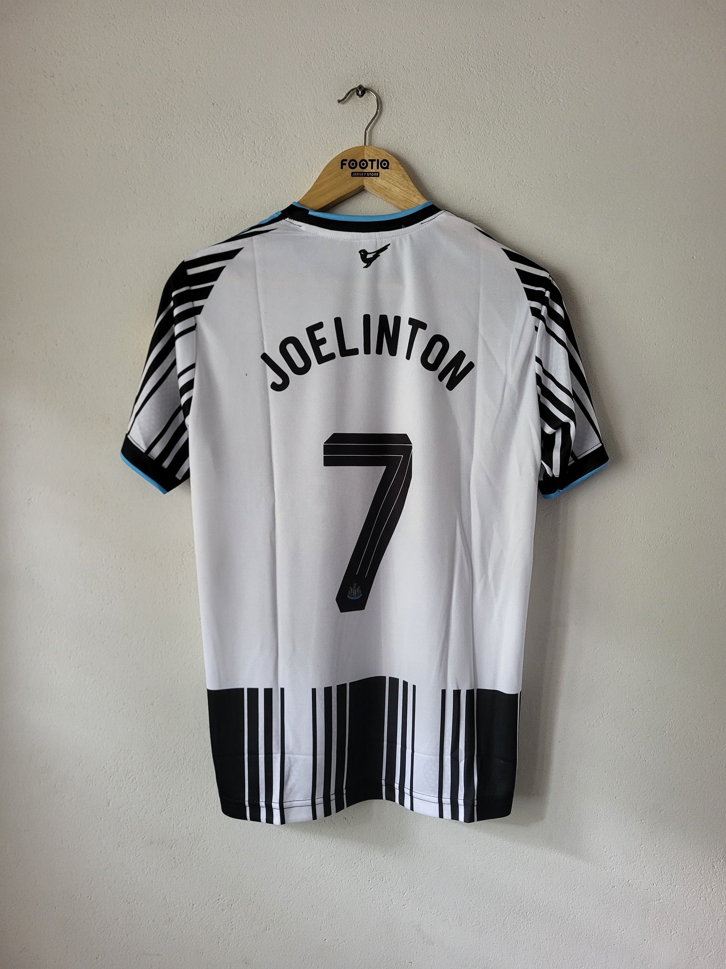A white soccer jersey with 'JOELINTON' and the number '7' printed on the back, hanging on a wooden hanger against a plain wall. The jersey features black and white striped patterns on the sleeves and hem.