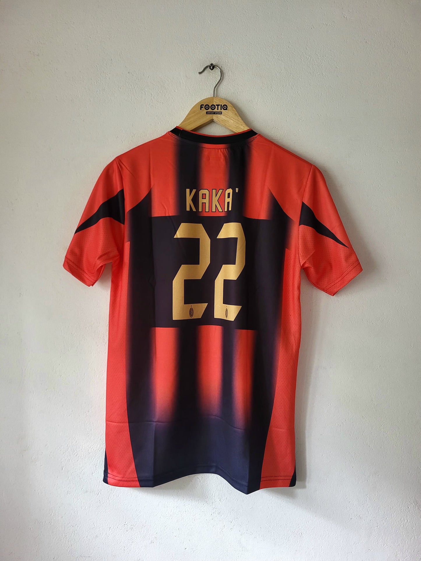 A red and navy blue striped soccer jersey with gold lettering and numbers, hanging on a wooden hanger against a plain wall. The back of the shirt features the name 'KAKA' and the number '22' in gold, and the hanger has 'FOXTROT' printed on it.