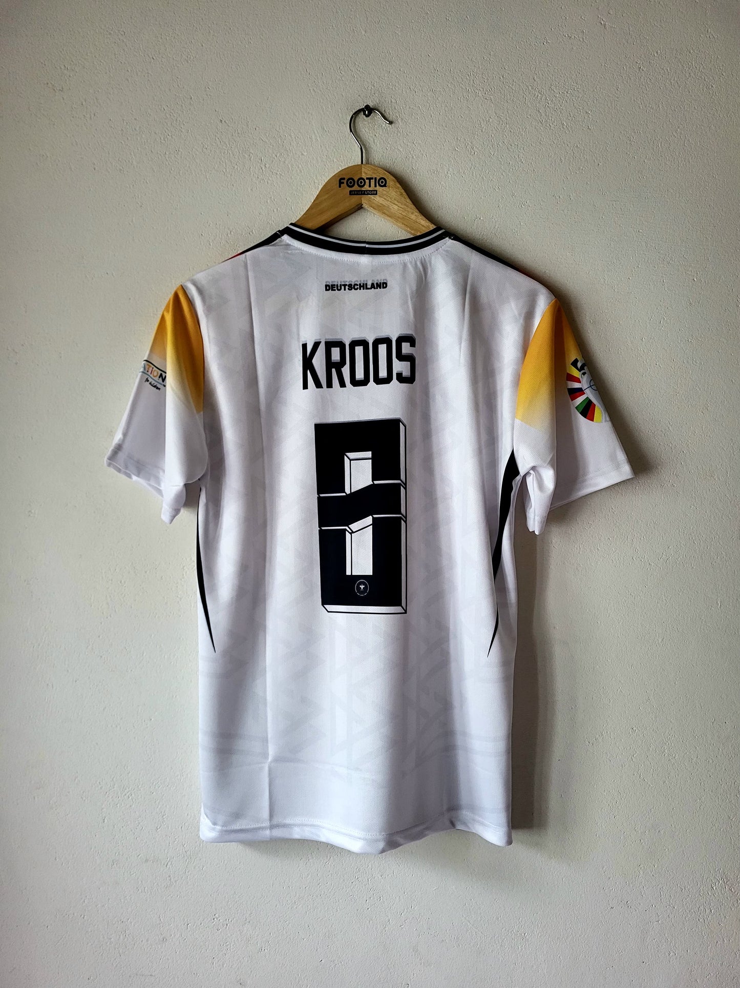 A white short-sleeve soccer jersey with 'KROOS' and the number 9 printed on the back, hanging on a wooden hanger against a light beige wall.