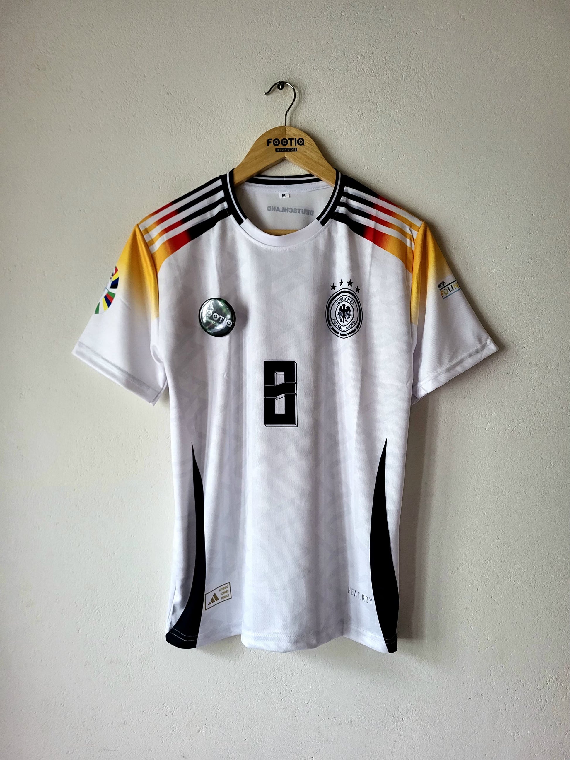 A white Germany national football team jersey with black and yellow accents, hanging on a wooden hanger against a plain beige wall.