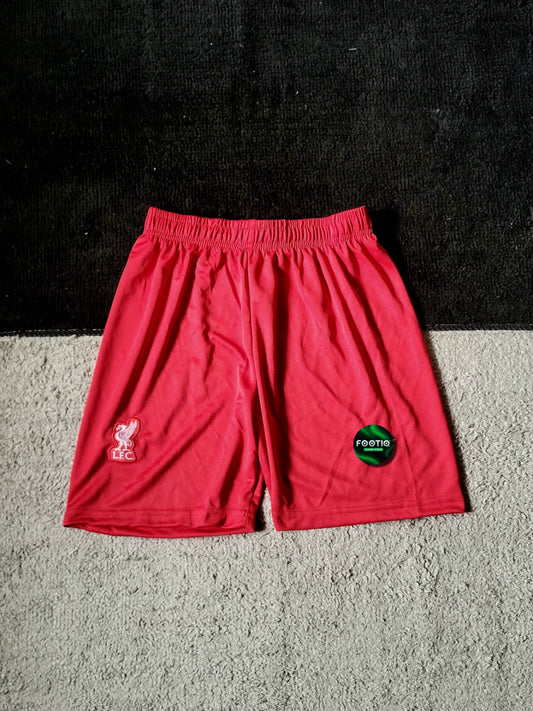 Red athletic shorts with an elastic waistband, featuring a white Liverpool FC crest on the left leg and a green circular 'foamit' logo on the right leg, laid flat on a textured gray carpet.