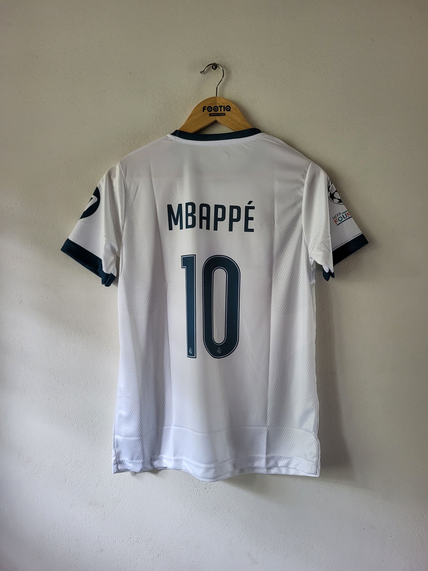 White soccer jersey with 'MBAPPÉ 10' printed on the back, hanging on a wooden hanger against a plain wall.
