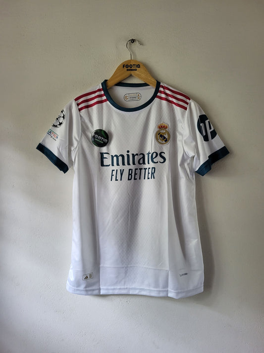 A white Real Madrid soccer jersey with navy and red accents, hanging on a wooden hanger against a plain beige wall. The jersey features the Emirates Fly Better sponsor logo, the Real Madrid crest, and the Adidas logo.