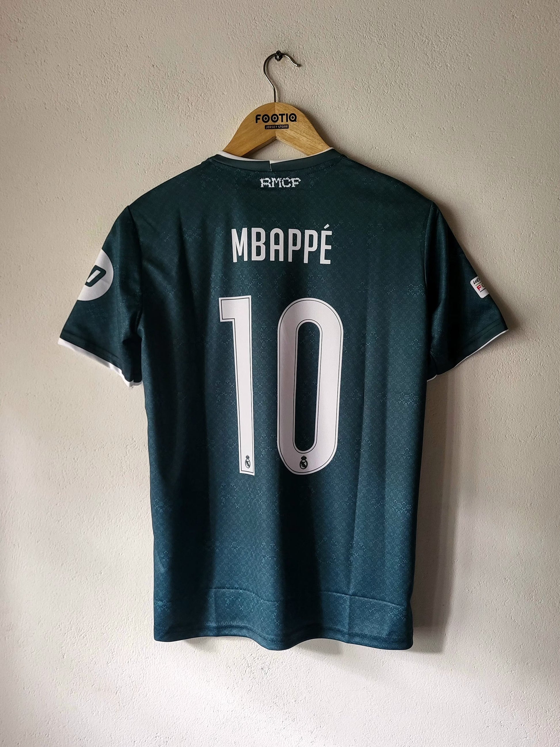 Back view of a dark teal RMC CP Mbappé jersey with the number 10 and name printed in white, hanging on a wooden hanger against a light beige wall.