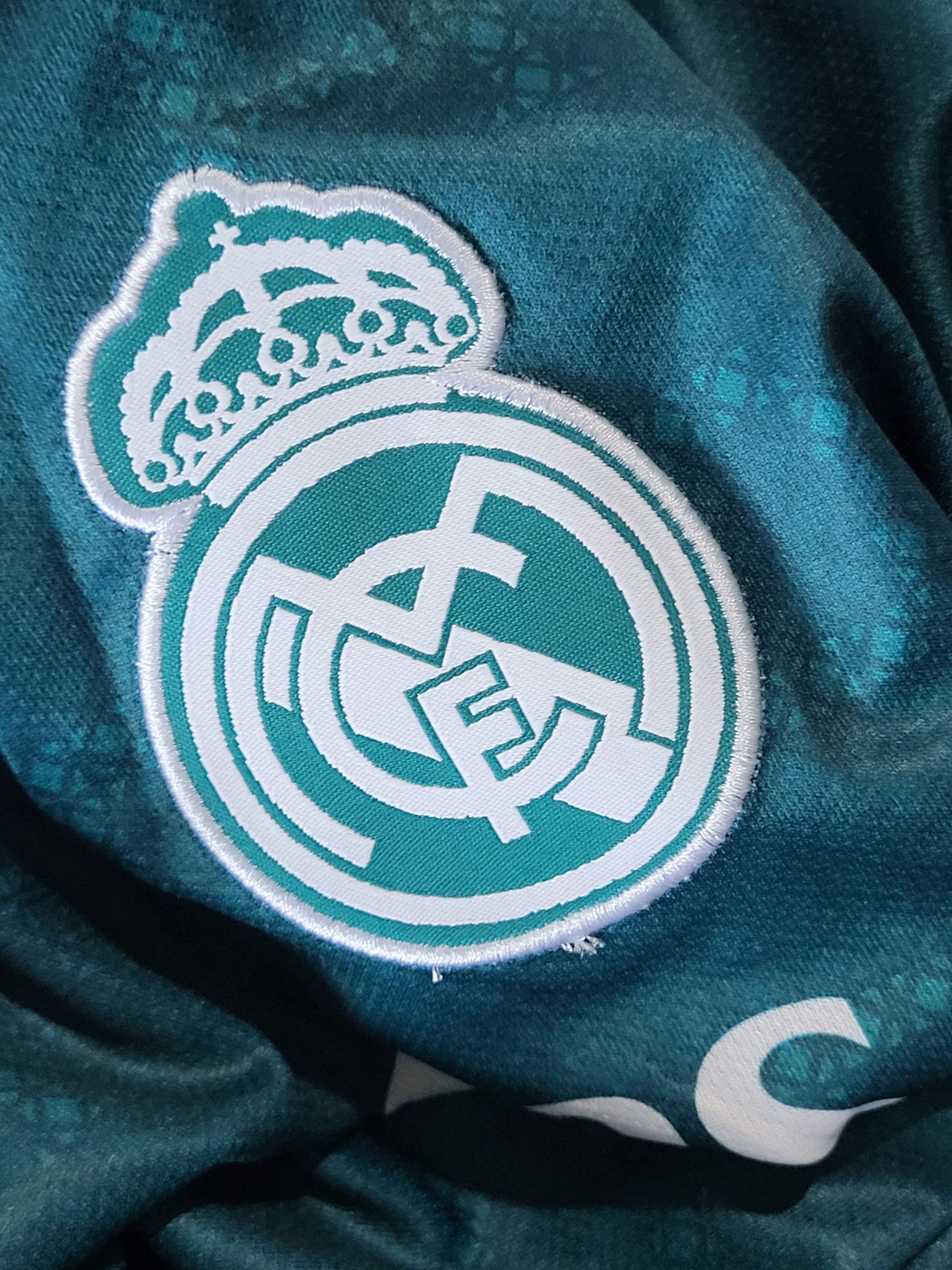 Close-up of a Real Madrid embroidered crest patch on a teal soccer jersey fabric.