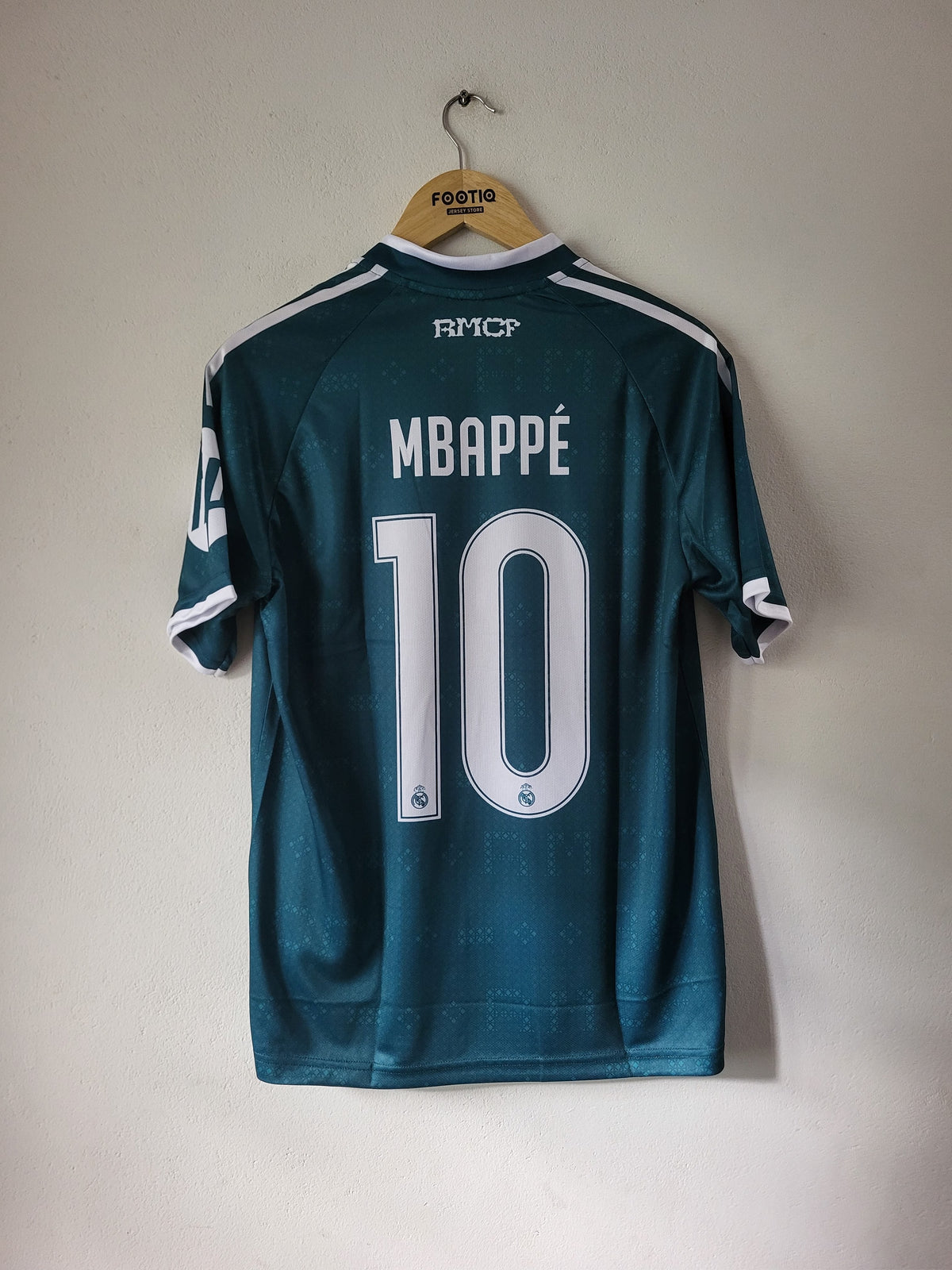 A dark teal FC Copenhagen soccer jersey with 'MBAPPÉ 10' printed on the back, hanging on a wooden hanger against a plain off-white wall.