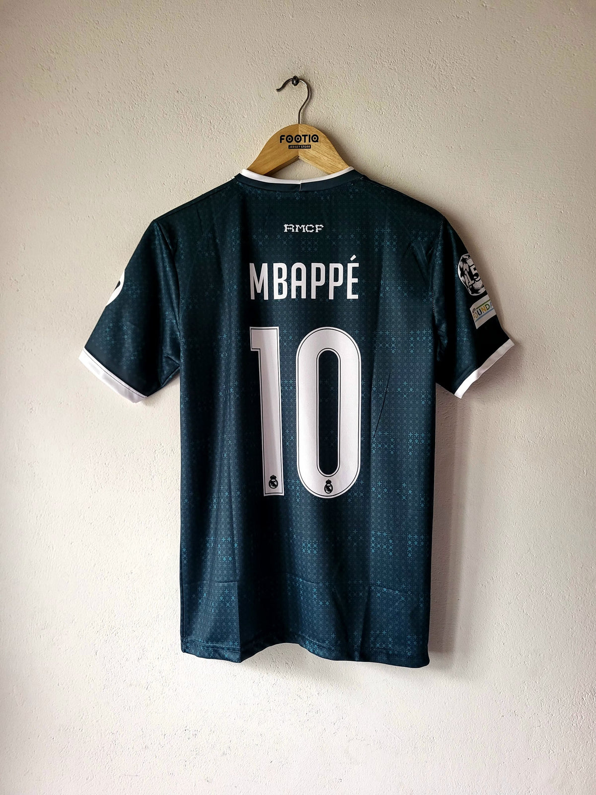 A dark teal Real Madrid soccer jersey with 'MBAPPÉ 10' printed on the back, hanging on a wooden hanger against a textured off-white wall.