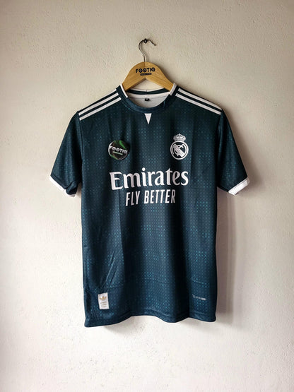 A dark teal Real Madrid soccer jersey hanging on a wooden hanger against a textured off-white wall. The jersey features white shoulder stripes, the Real Madrid crest, a green sponsor patch, and 'Emirates FLY BETTER' printed in white across the chest.