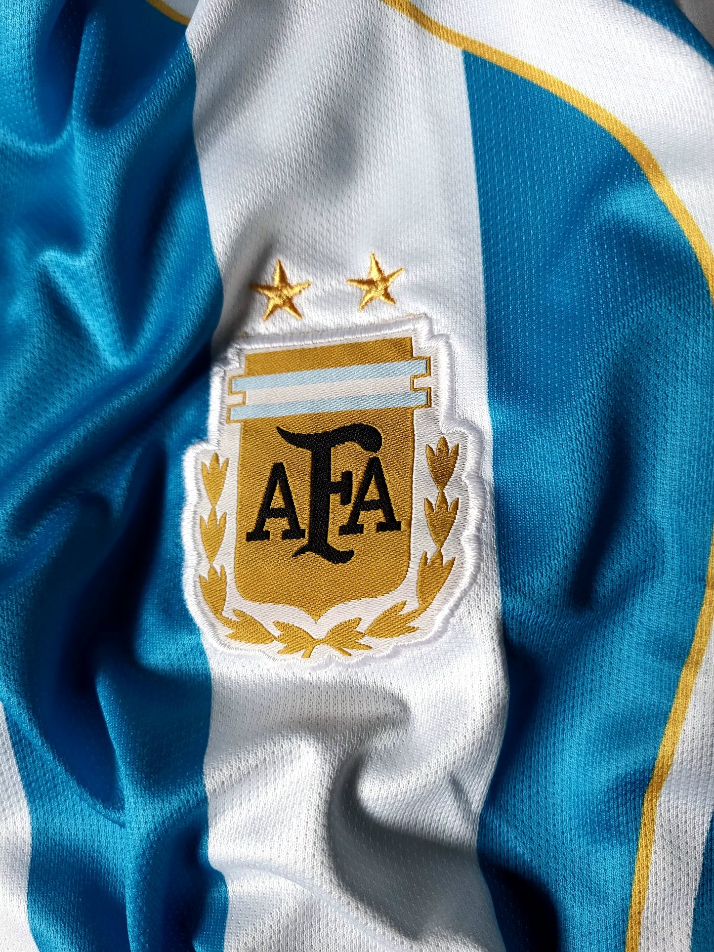 Close-up of an Argentina national football team jersey showing the blue and white vertical stripes with gold trim and the embroidered AFA crest with two gold stars.