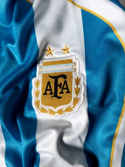 Close-up of an Argentina national football team jersey showing the blue and white vertical stripes with gold trim and the embroidered AFA crest with two gold stars.