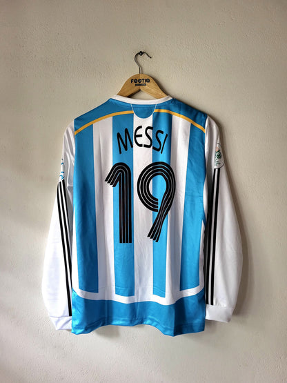 A long-sleeve Argentina soccer jersey with light blue and white vertical stripes, featuring 'MESSI' and the number '19' on the back. The jersey is hanging on a wooden hanger with 'FOSTIE' branding, against a plain beige wall.