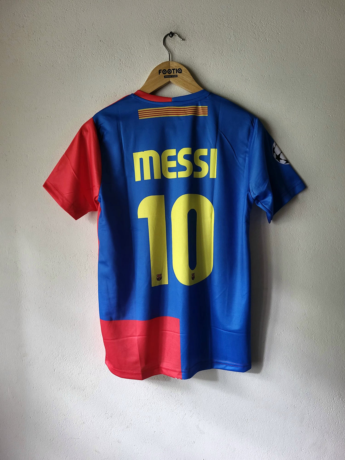 A blue and red soccer jersey with yellow 'MESSI 10' lettering on the back, hanging on a wooden hanger against a white wall.