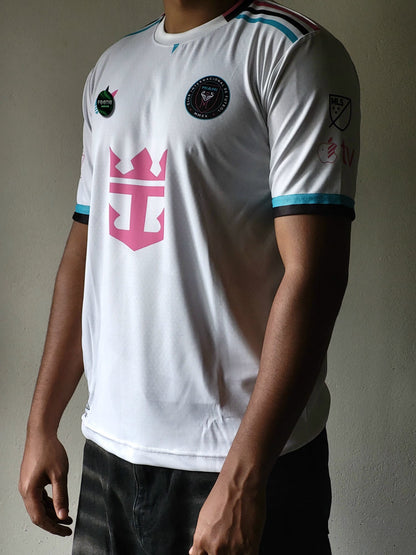 A white short-sleeve soccer jersey with a large pink crown-and-anchor style logo on the chest, black and light blue trim, and multiple circular patches, shown on a cropped male model against a plain wall.