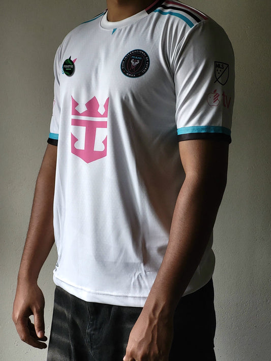 A white short-sleeve soccer jersey with a large pink crown-and-anchor style logo on the chest, black and light blue trim, and multiple circular patches, shown on a cropped male model against a plain wall.