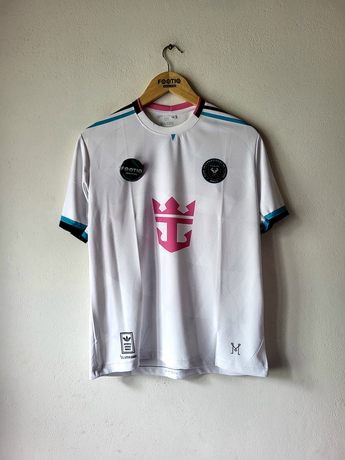 A white short-sleeve soccer jersey hanging on a wooden hanger against a textured off-white wall. The jersey features a pink and black crown logo on the center chest, black circular patches on the shoulders, and teal and black striped accents on the shoulders and sleeve cuffs.