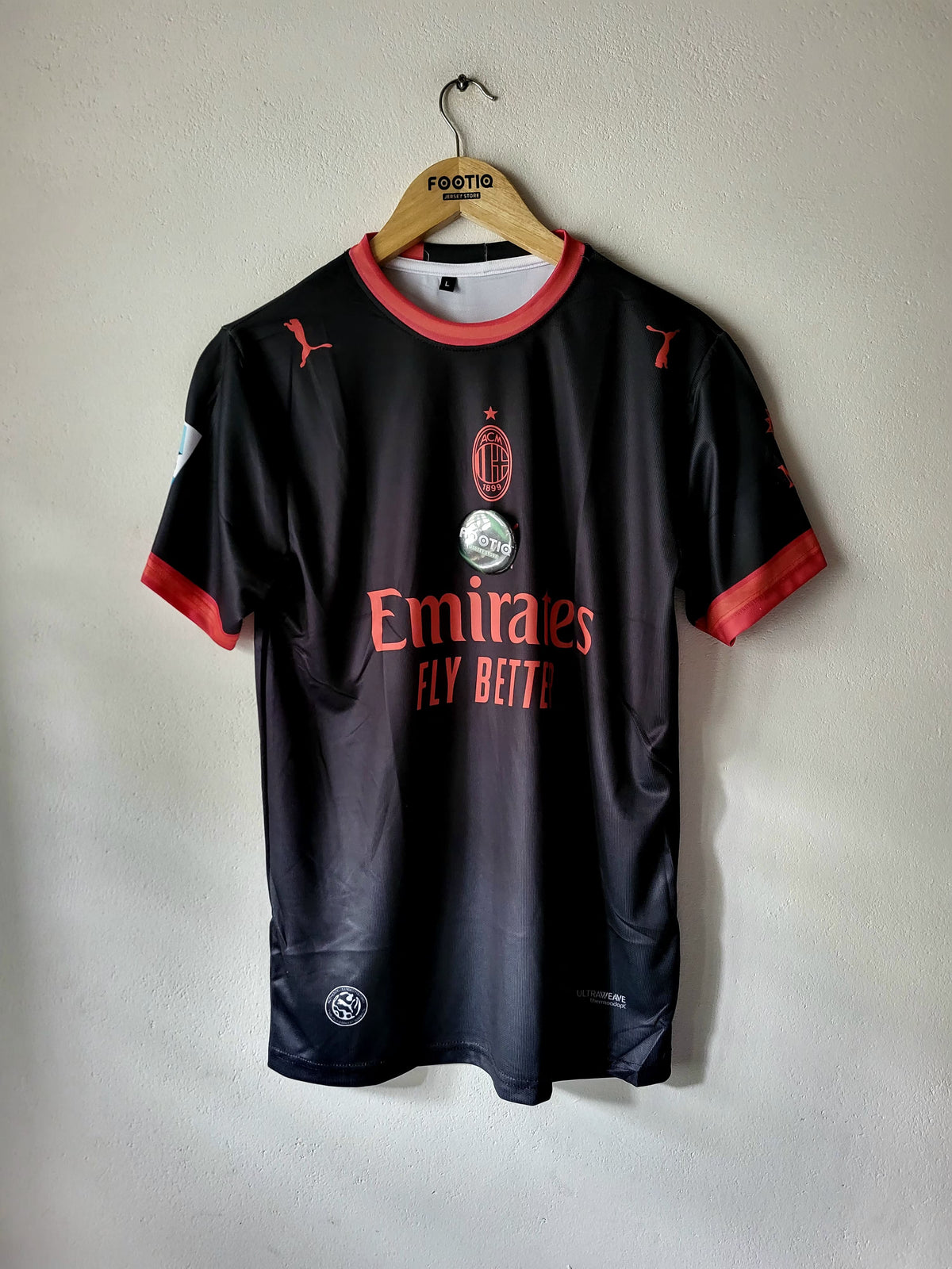 A black AC Milan soccer jersey with red trim, hanging on a wooden hanger against a light gray wall. The jersey features the AC Milan crest, the Emirates Fly Beite sponsor logo, and Puma branding.