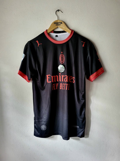 A black AC Milan soccer jersey with red trim, hanging on a wooden hanger against a light gray wall. The jersey features the AC Milan crest, the Emirates Fly Beite sponsor logo, and Puma branding.
