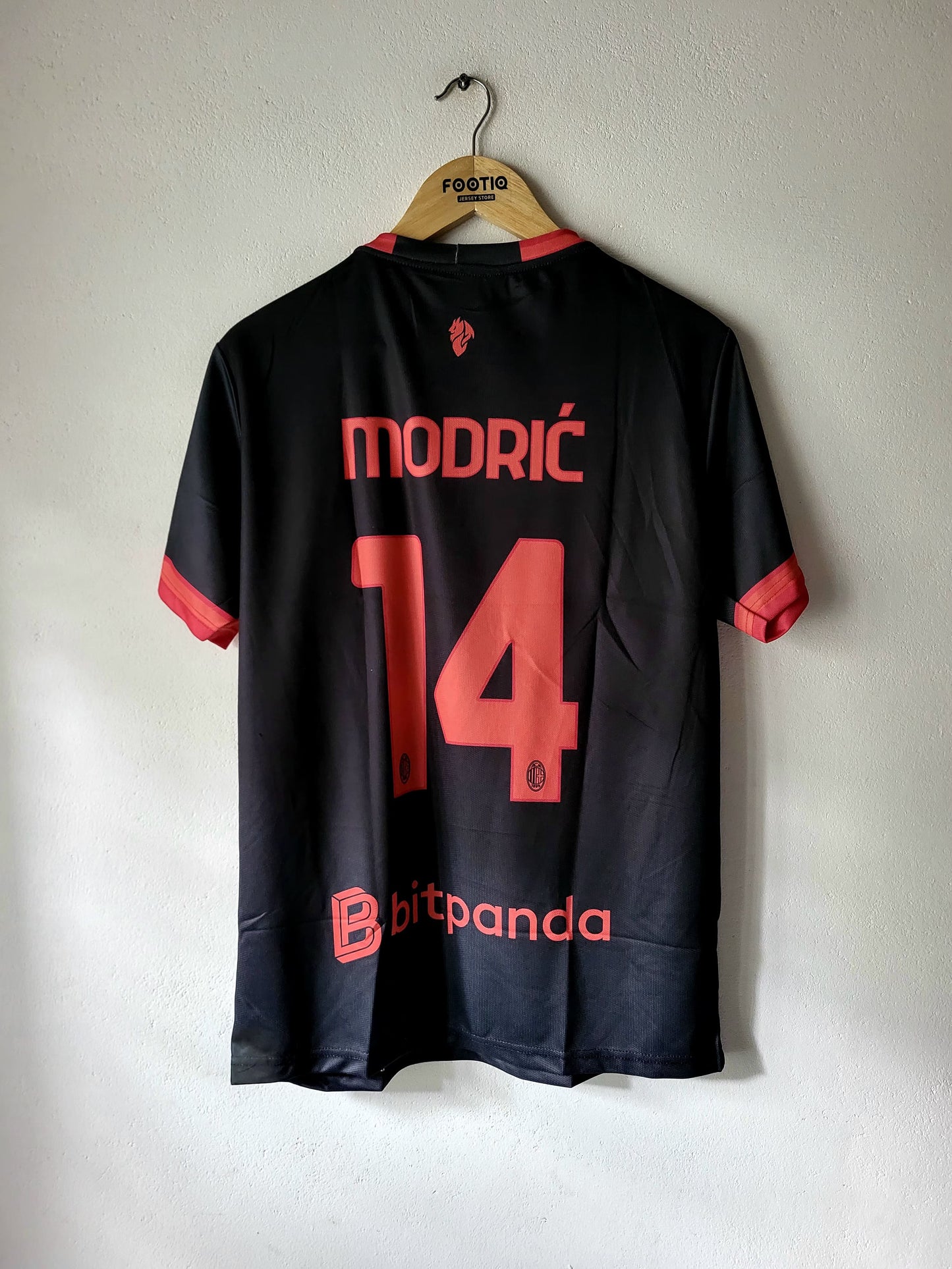 A black soccer jersey with red accents hanging on a wooden hanger against a white wall. The back of the shirt features the name 'MODRIČ' and the number '14' in red, with a 'bitpanda' sponsor logo near the bottom.
