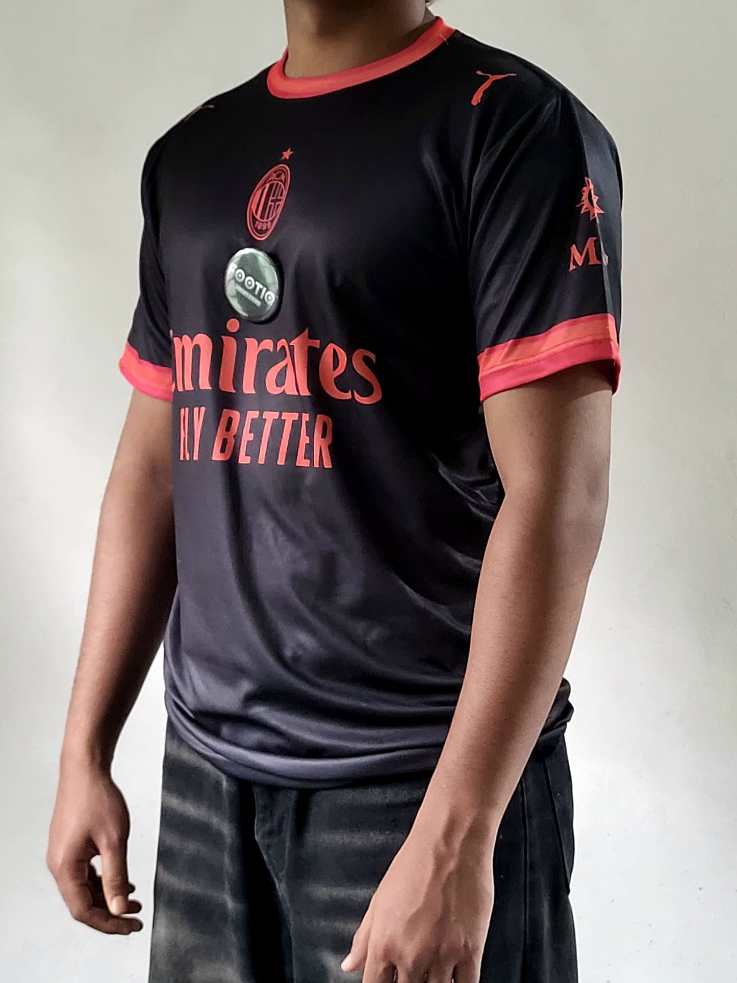A person wearing a black AC Milan soccer jersey with red trim and red text, paired with dark jeans, photographed against a plain light gray background.