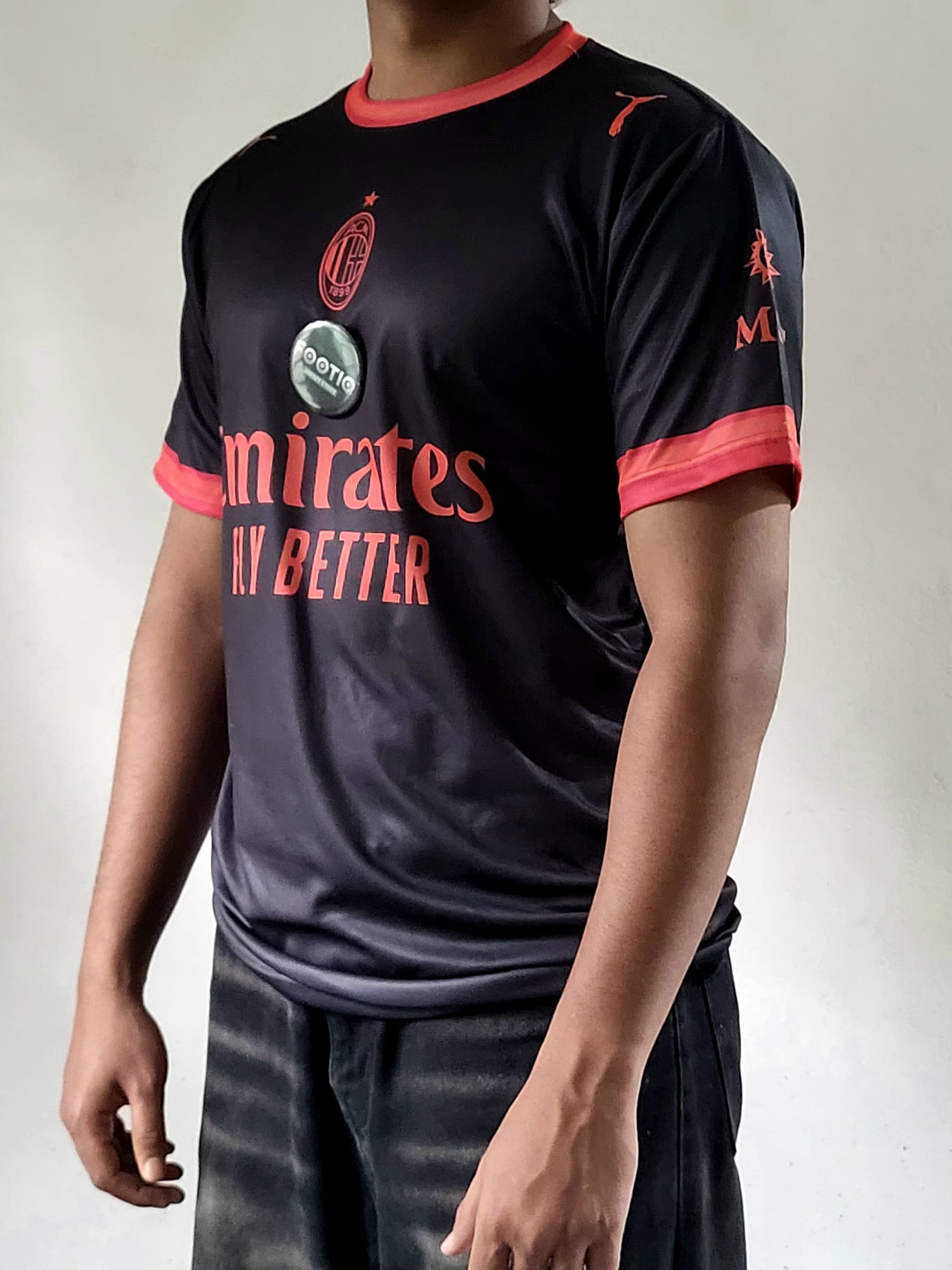 A person wearing a black AC Milan soccer jersey with red trim and red text, paired with dark jeans, photographed against a plain light gray background.