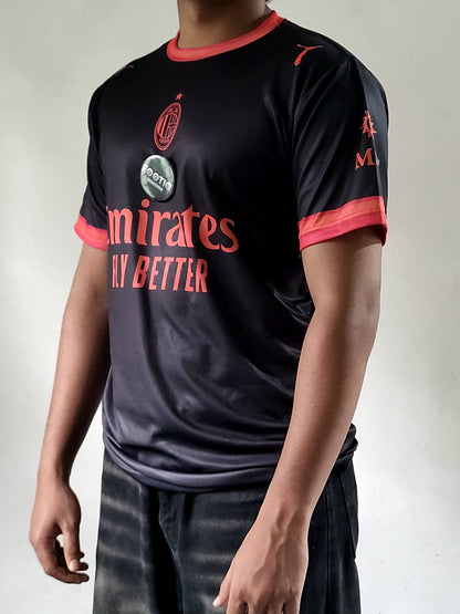 A person wearing a black AC Milan soccer jersey with red trim and red text, paired with dark jeans, photographed against a plain light gray background.