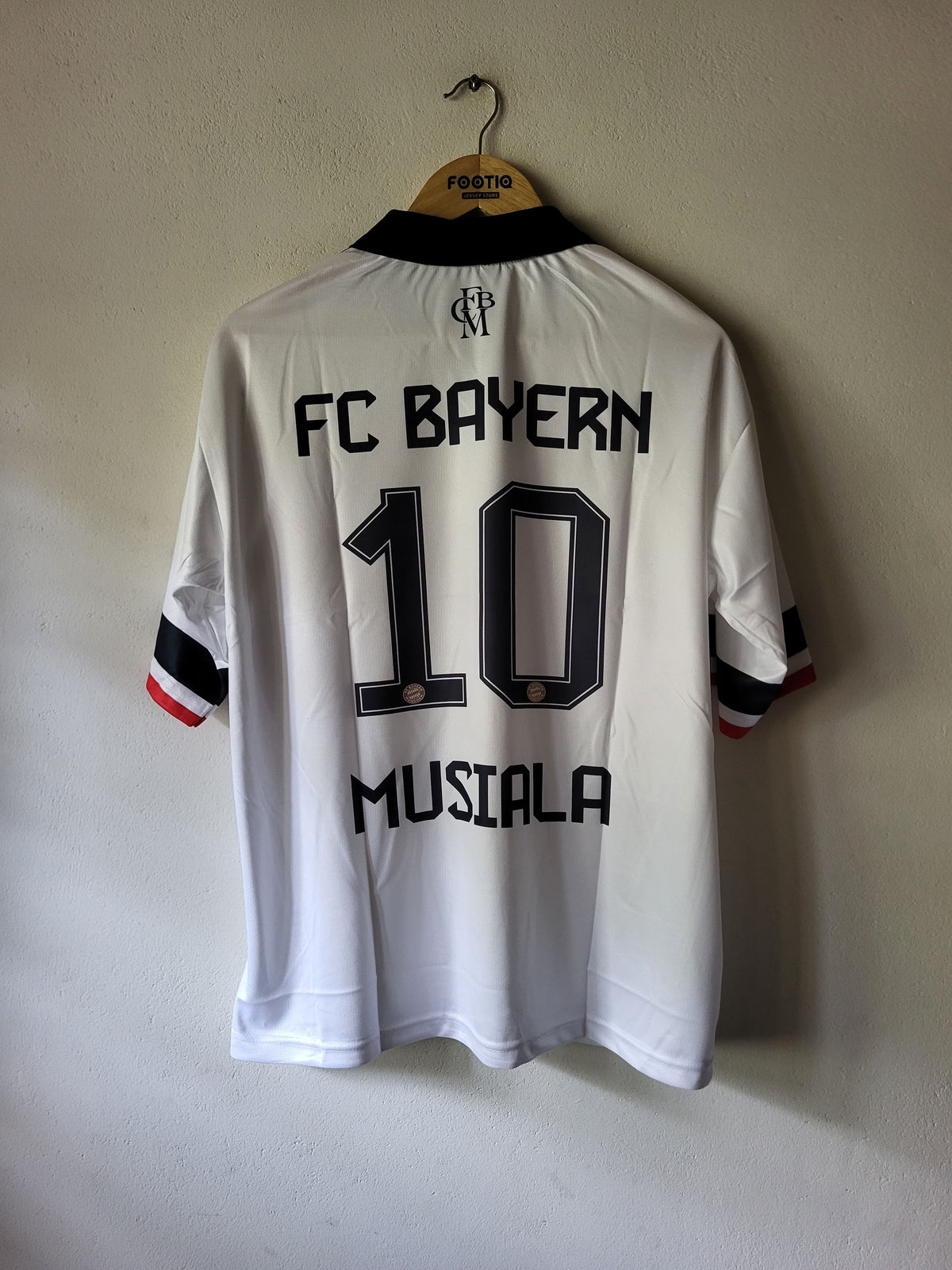 White FC Bayern Munich 10 number back jersey with Musiala name and crest, hanging on a wooden hanger against a plain wall.