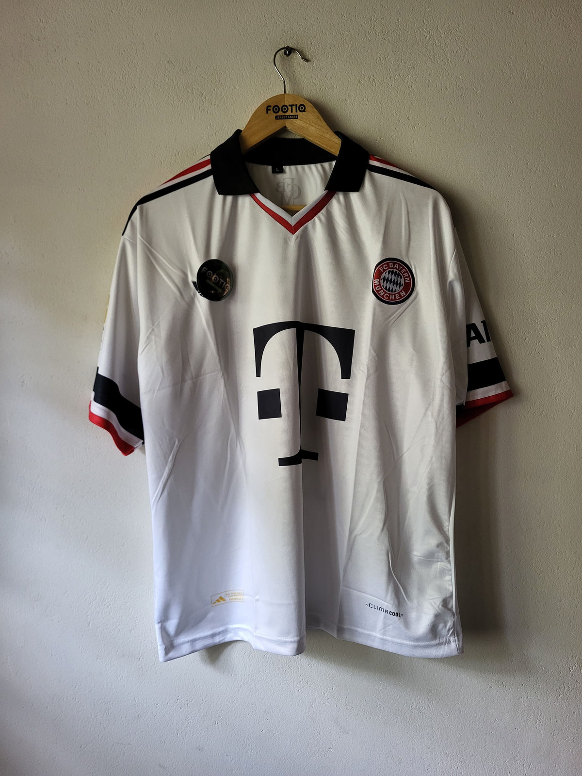 A vintage white Bayern Munich soccer jersey with black and red accents, hanging on a wooden hanger against a plain wall. The jersey features the large black T-Mobile logo on the front, the Bayern Munich club crest on the left chest, and a Festig brand label on the hanger.
