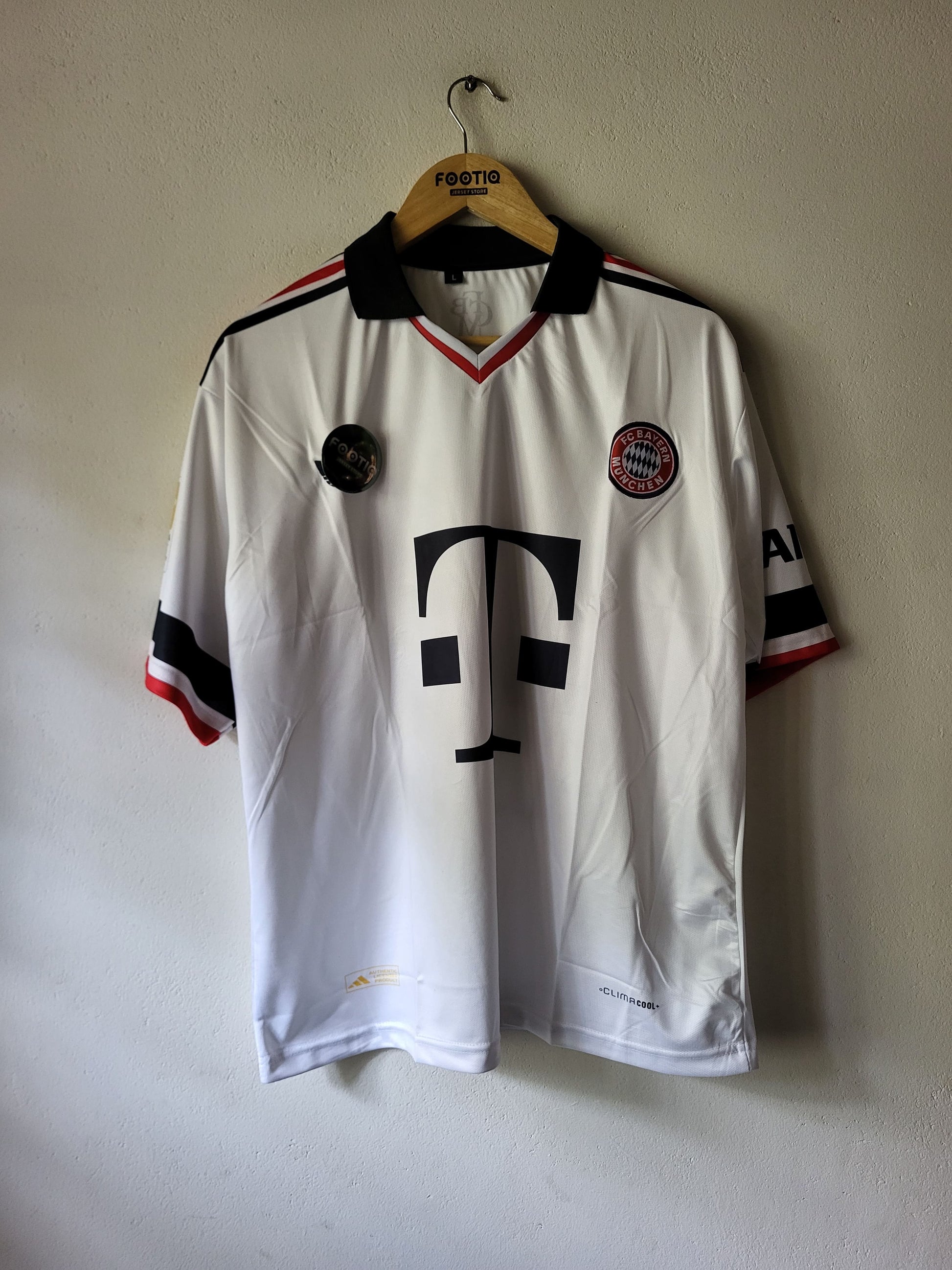 A vintage white Bayern Munich soccer jersey with black and red accents, hanging on a wooden hanger against a plain wall. The jersey features the large black T-Mobile logo on the front, the Bayern Munich club crest on the left chest, and a Festig brand label on the hanger.