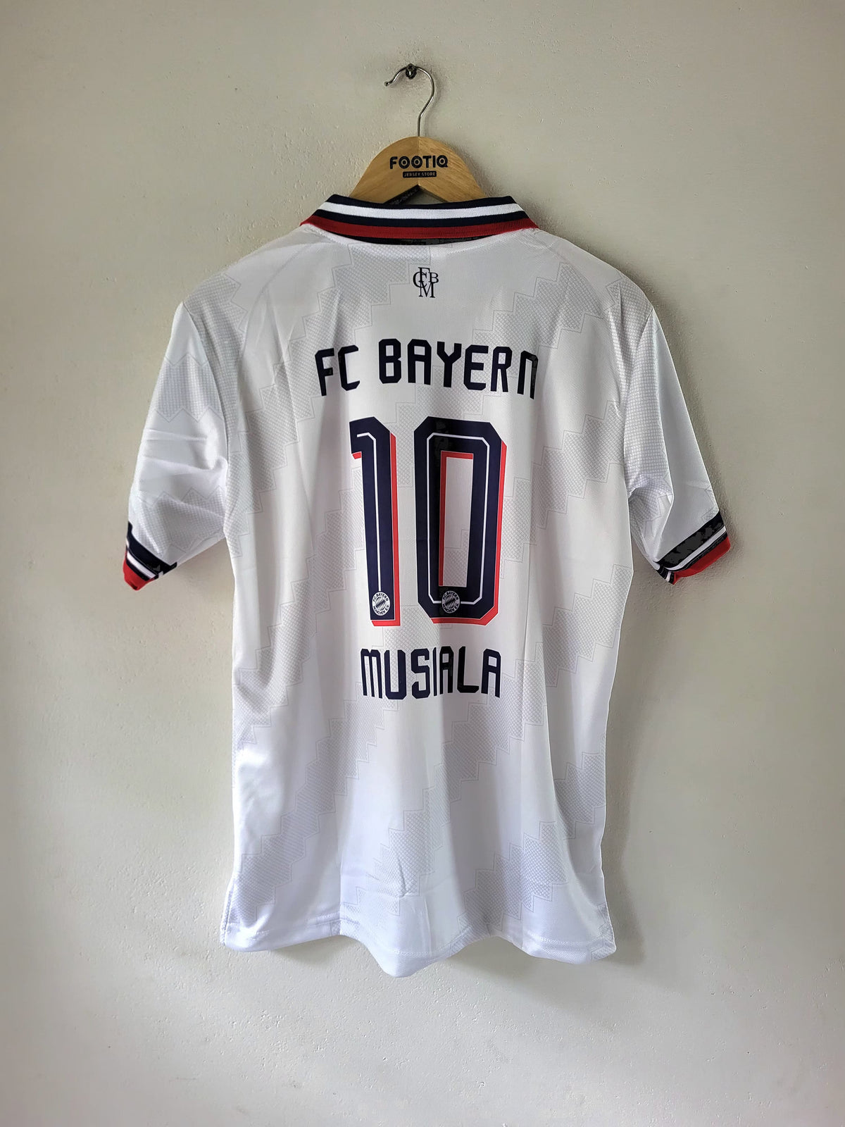 A white FC Bayern Munich short-sleeve soccer jersey with '10 MUSIALA' printed on the back, hanging on a wooden hanger against a plain beige wall.