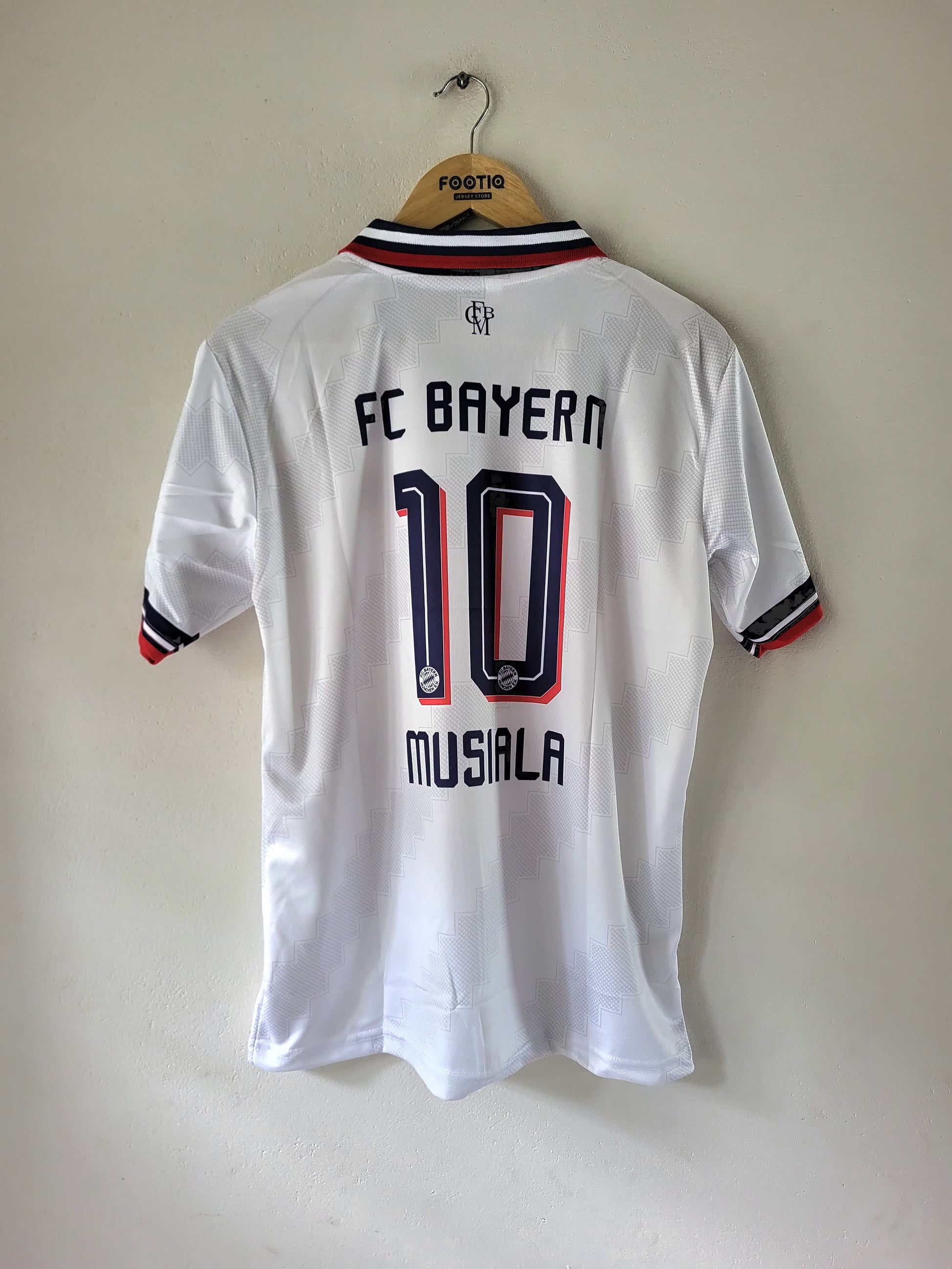 A white FC Bayern Munich short-sleeve soccer jersey with '10 MUSIALA' printed on the back, hanging on a wooden hanger against a plain beige wall.
