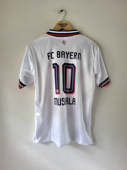 A white FC Bayern Munich short-sleeve soccer jersey with '10 MUSIALA' printed on the back, hanging on a wooden hanger against a plain beige wall.