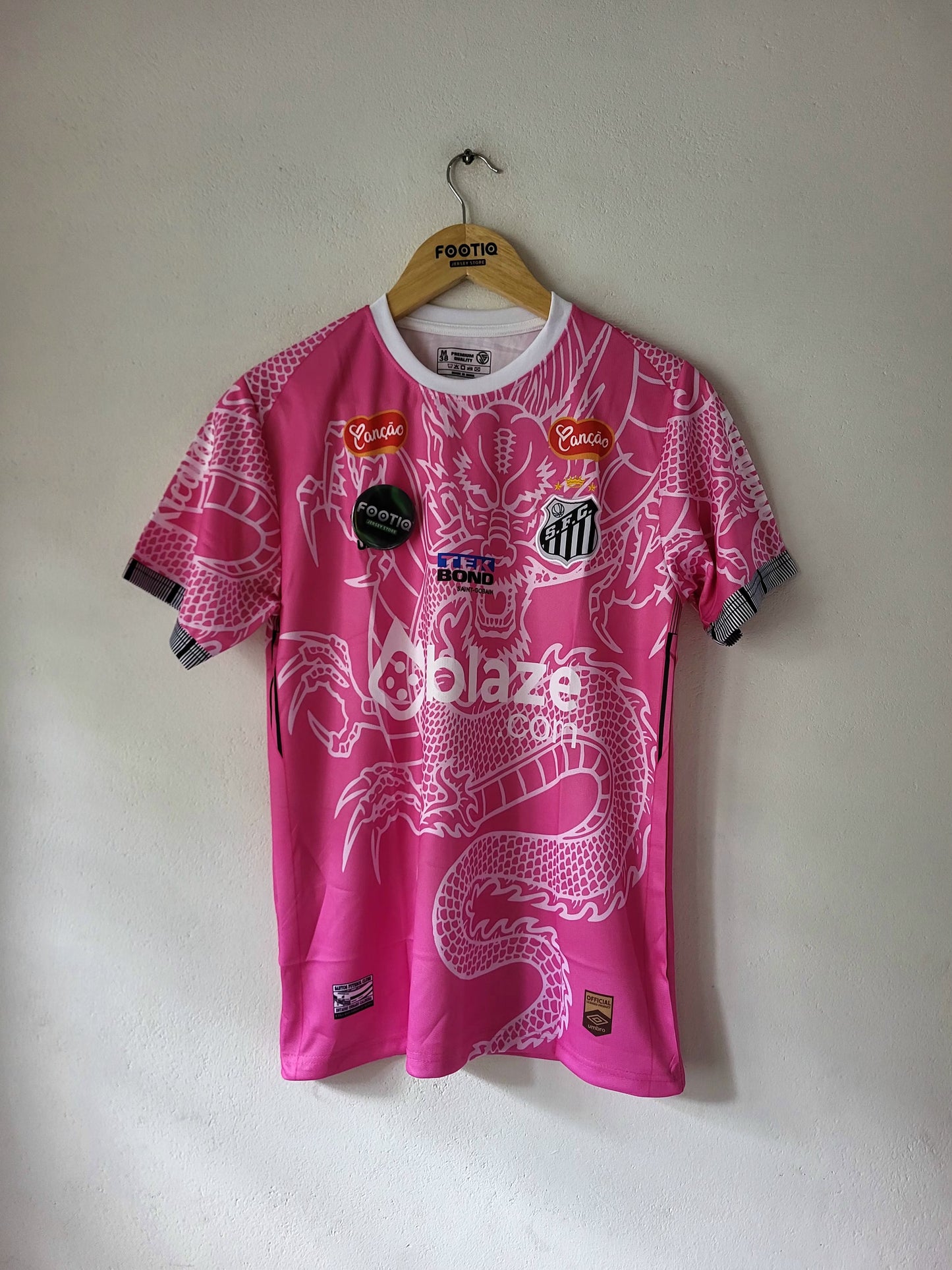 A pink and white soccer jersey with a dragon graphic, hanging on a wooden hanger against a plain wall. The jersey features multiple sponsor logos and a central crest.