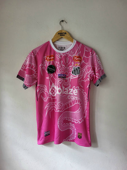 A pink and white soccer jersey with a dragon graphic, hanging on a wooden hanger against a plain wall. The jersey features multiple sponsor logos and a central crest.