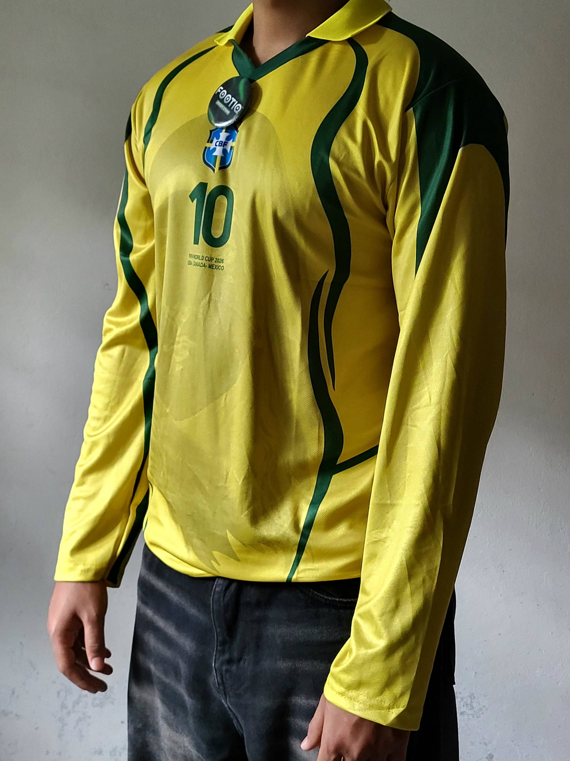 A person wearing a yellow long-sleeve soccer jersey with dark green shoulder panels and green curved stripe details, featuring the number 10 and a Brazil crest. The image is cropped from the neck down, showing the person's hands and dark jeans against a gray textured wall.