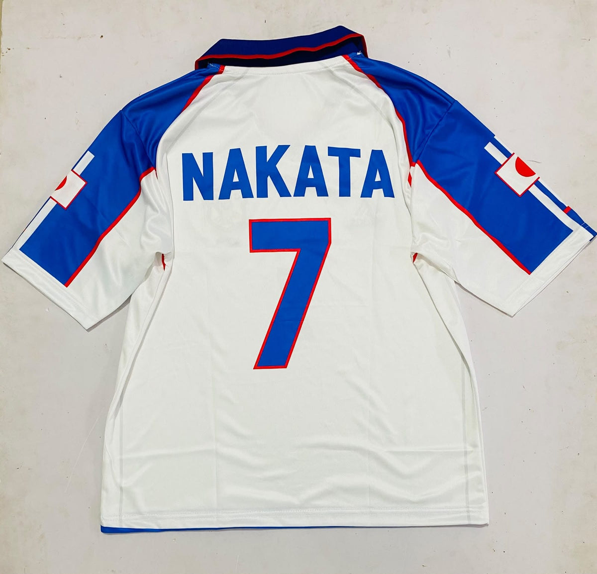Back view of a white soccer jersey with blue and red trim, featuring the name 'NAKATA' and number '7' in blue with red outlines on the back.