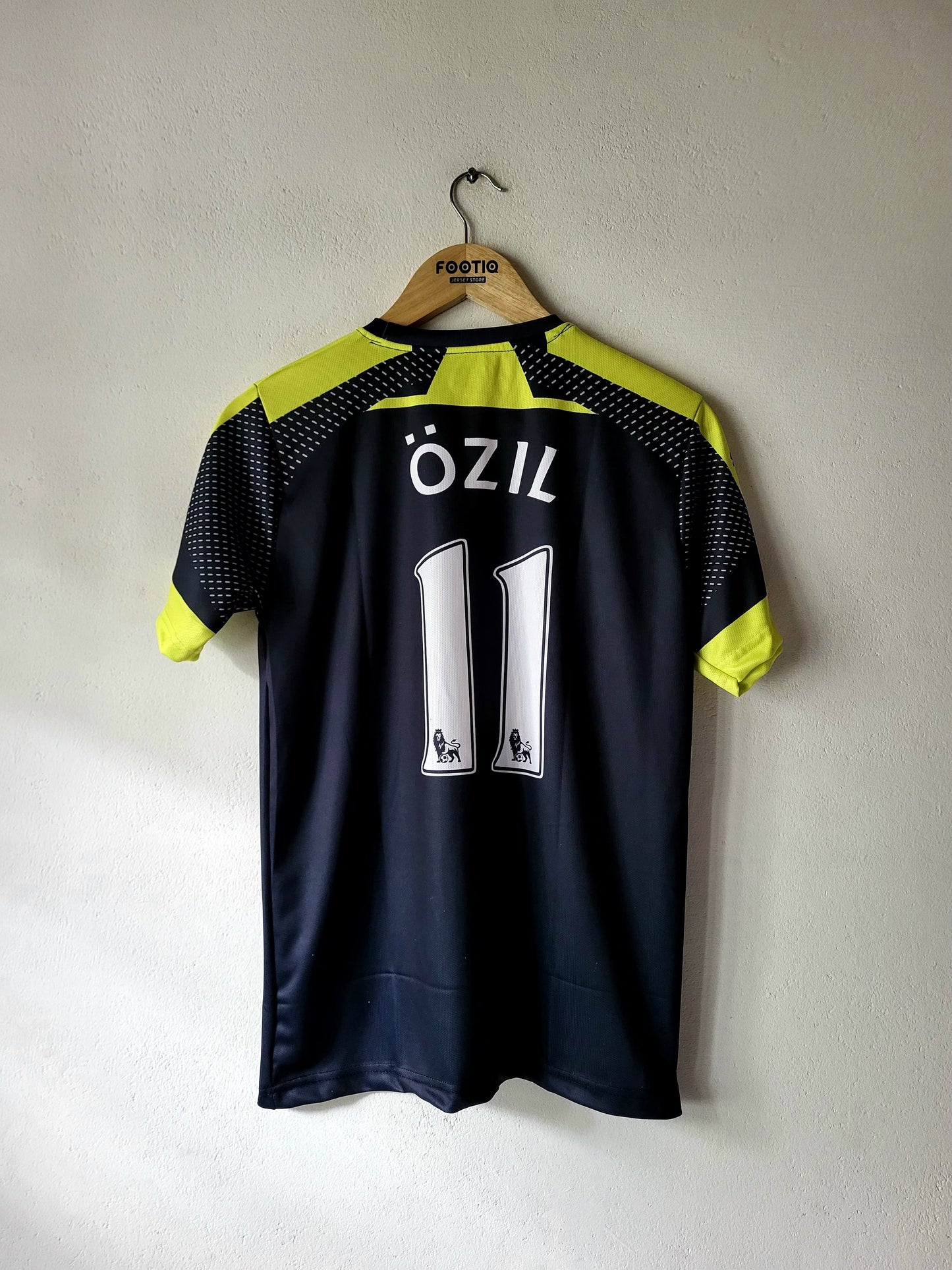 A dark navy blue soccer jersey with neon yellow-green accents on the shoulders and sleeves, featuring the name 'ÖZIL' and number '11' on the back. The jersey is hanging on a wooden hanger with 'FOOTBALL' printed on it, against a plain light gray wall.