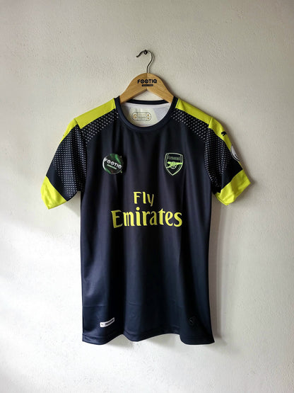 A dark navy blue Arsenal football jersey with neon yellow accents on the shoulders and sleeves, hanging on a wooden hanger against a plain white wall. The jersey features the 'Fly Emirates' sponsor logo on the front, along with the Arsenal and Puma branding.