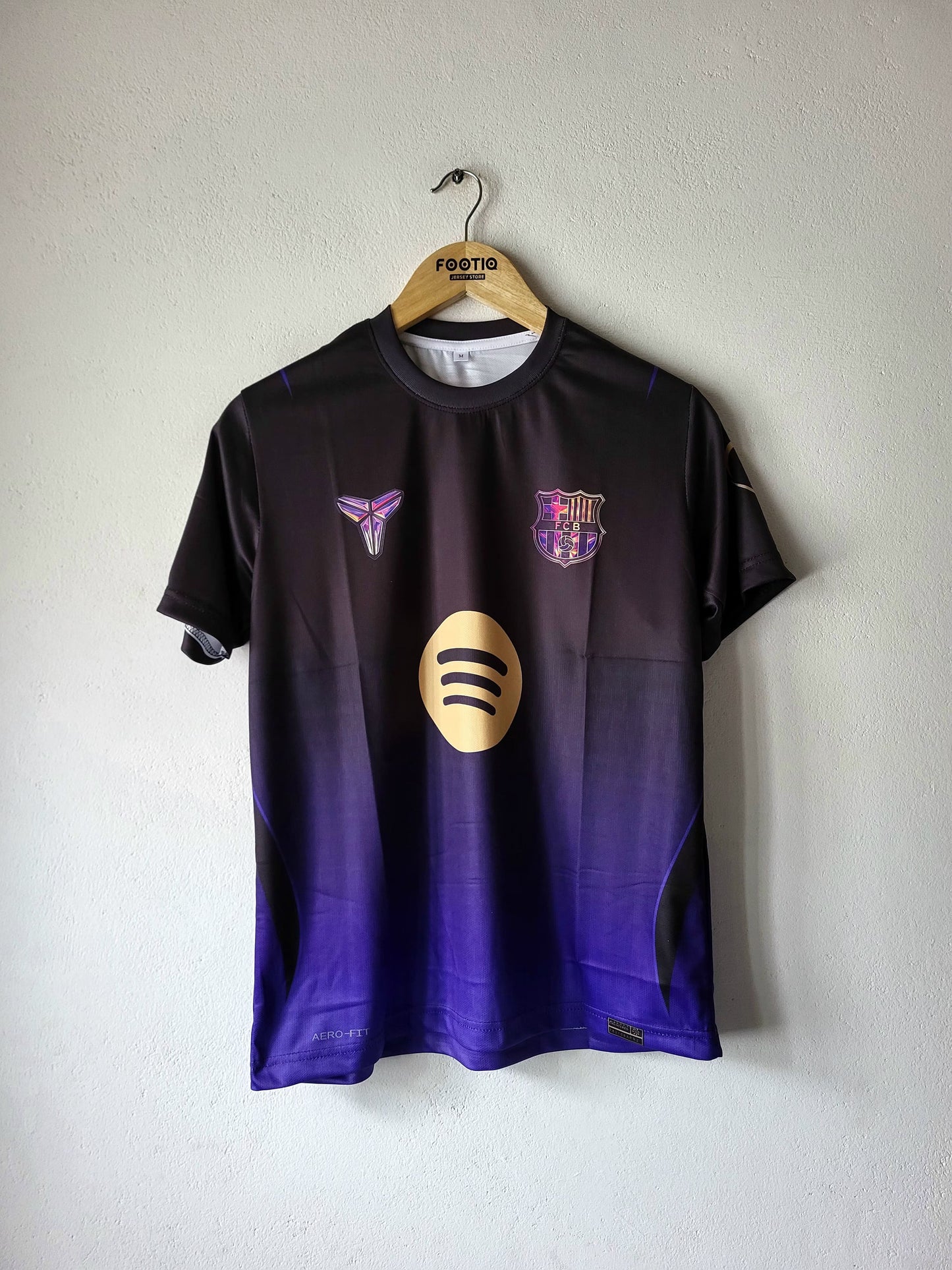A short-sleeve FC Barcelona soccer jersey hanging on a wooden hanger against a textured white wall. The jersey features a dark gradient transitioning from black at the top to deep purple at the bottom, with a gold Spotify sponsor logo on the center chest, a purple and gold Nike Swoosh on the right chest, and the FC Barcelona crest on the left chest.