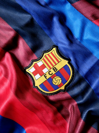 Close-up of an FC Barcelona soccer jersey fabric with the embroidered club crest centered on diagonal red, blue, and navy stripes.