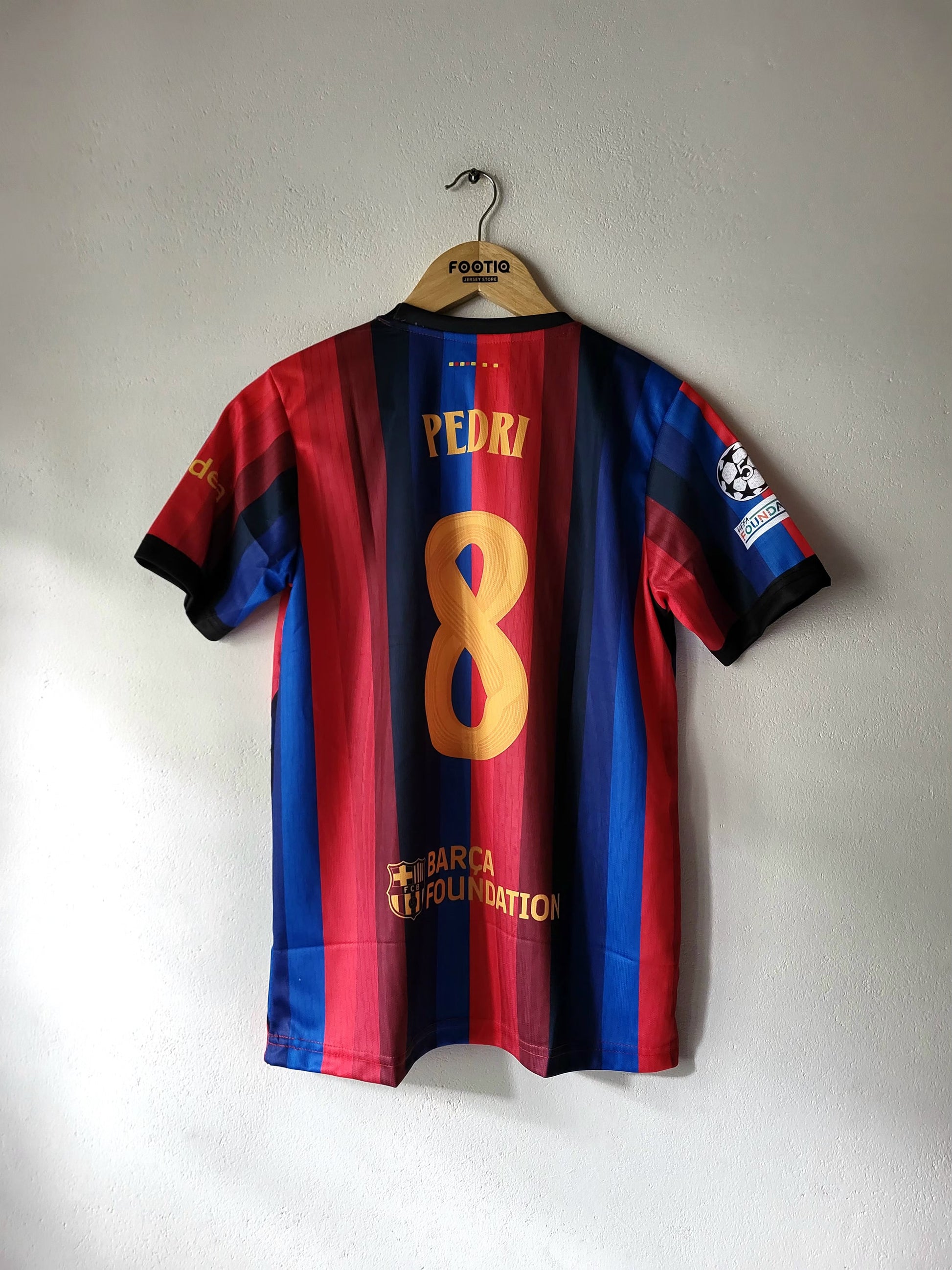A FC Barcelona home jersey with the name PEDRI and number 8 on the back, hanging on a wooden hanger against a plain wall.
