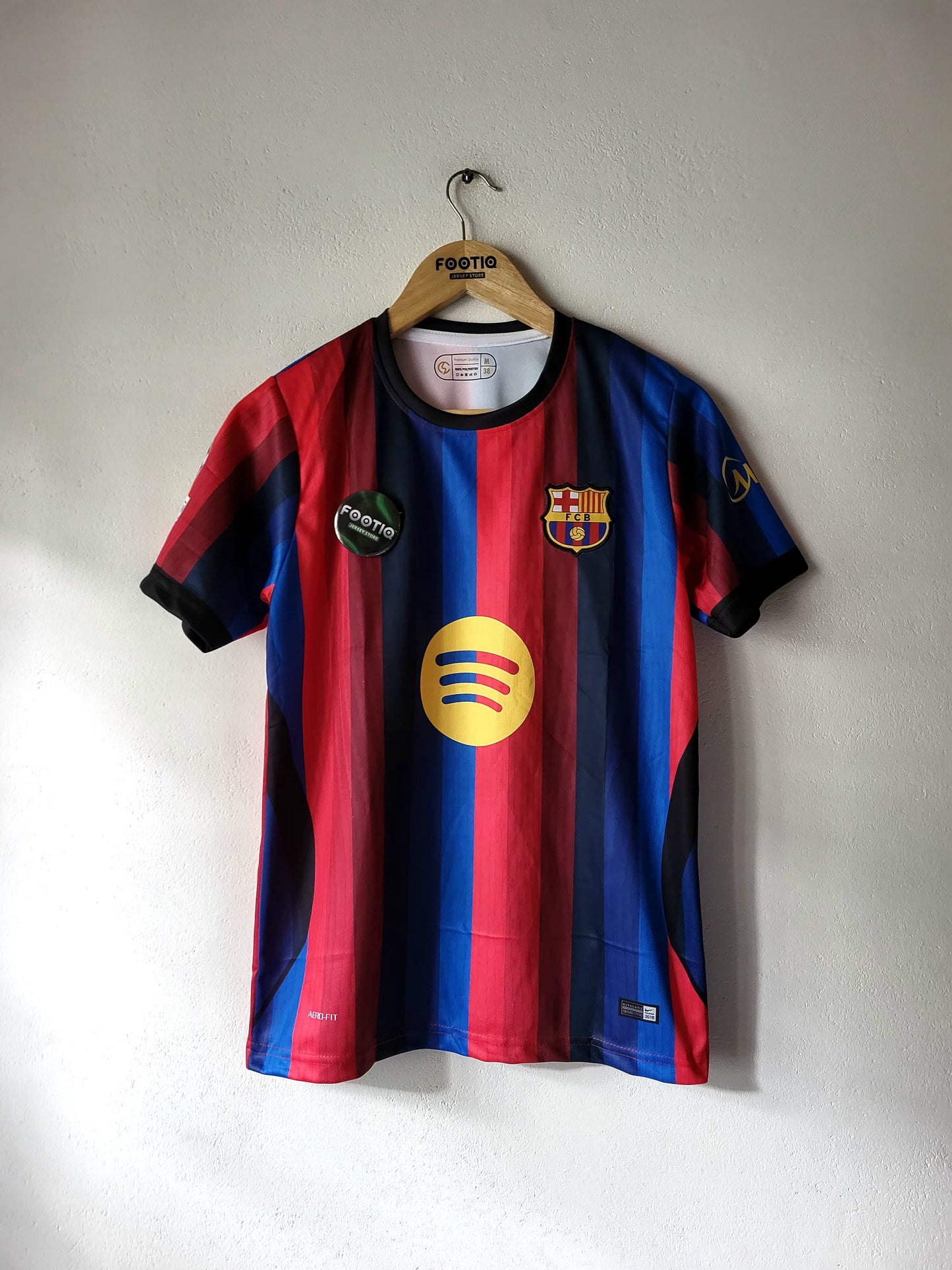 FC Barcelona home soccer jersey with red and blue stripes, hanging on a wooden hanger against a plain wall. Features the yellow Spotify sponsor logo on the chest.