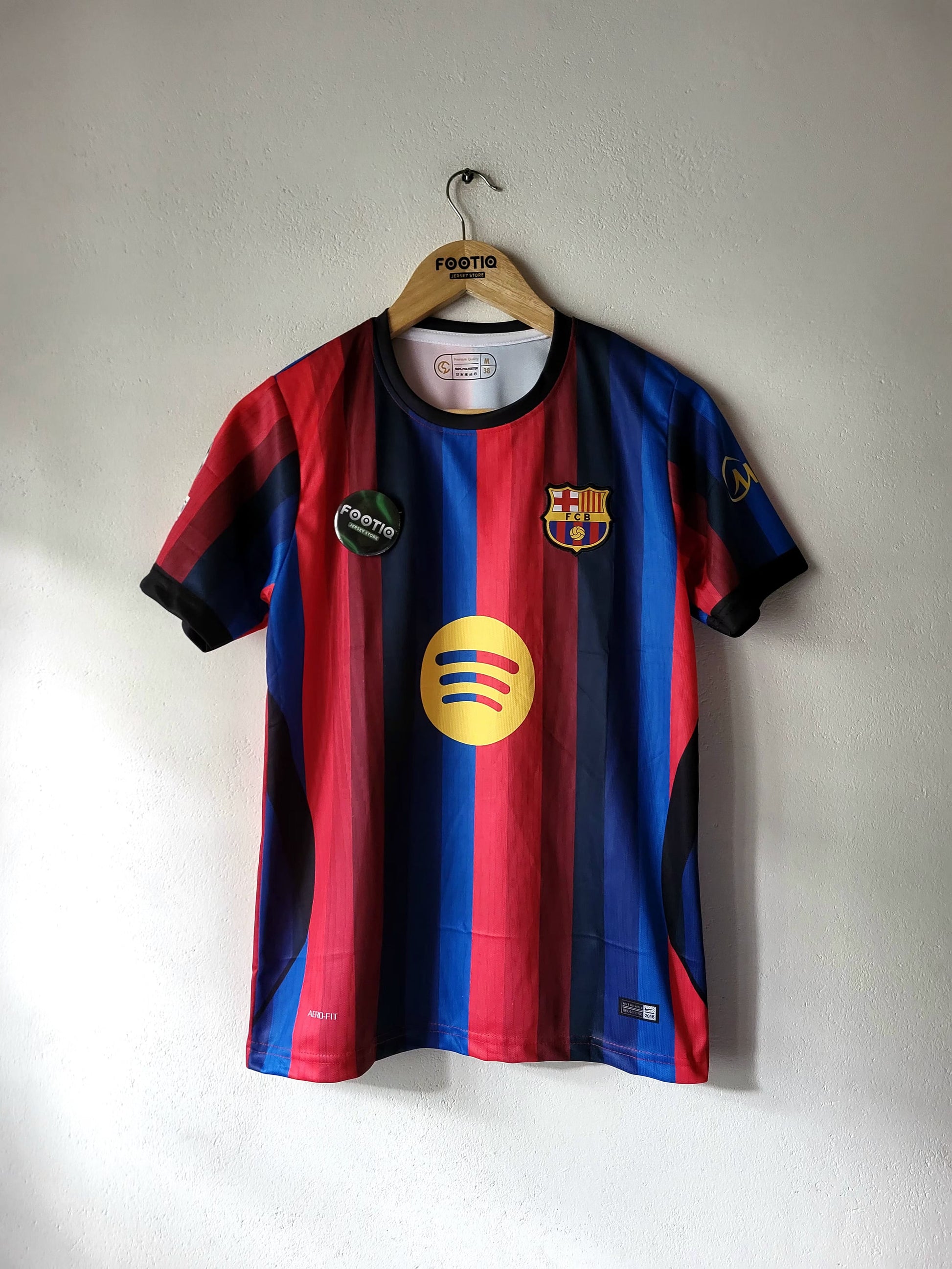 FC Barcelona home soccer jersey with red and blue stripes, hanging on a wooden hanger against a plain wall. Features the yellow Spotify sponsor logo on the chest.