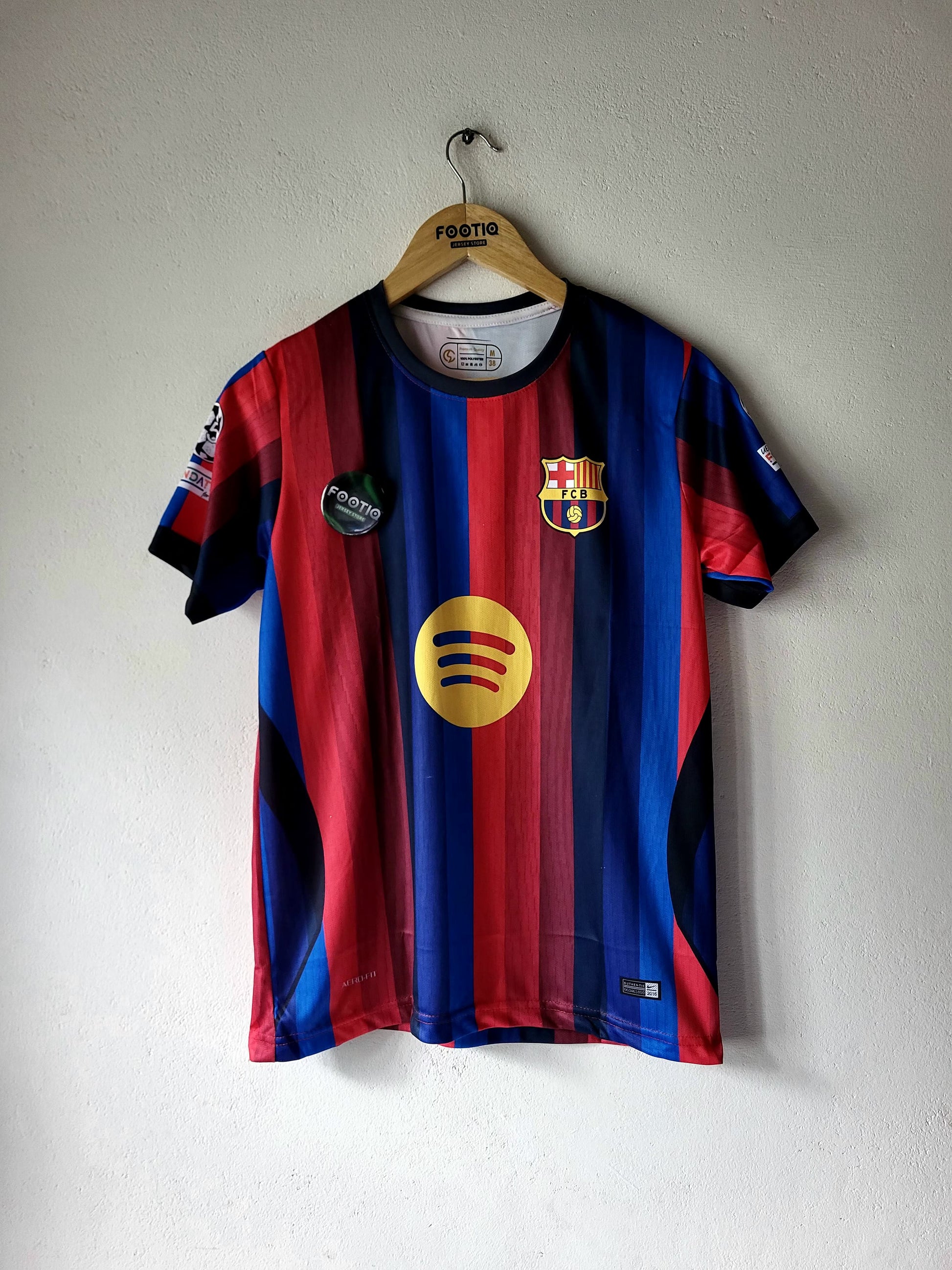 A short-sleeve FC Barcelona soccer jersey with red and blue vertical stripes, hanging on a wooden hanger against a white wall. The jersey features the club crest on the left chest, a yellow Spotify sponsor logo in the center, and a green 'Fiestas Barcelona' patch on the right chest.