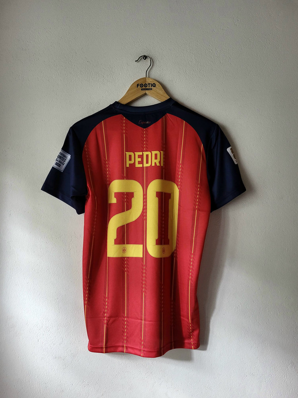 A red and navy blue soccer jersey with 'PEDRI' and the number '20' printed in yellow on the back, hanging on a wooden hanger against a plain wall.