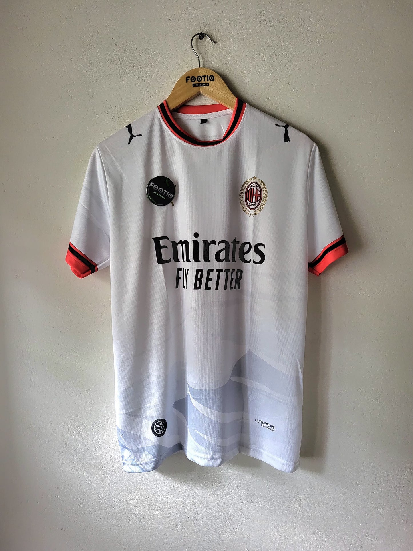 A white AC Milan soccer jersey with orange and black trim, hanging on a wooden hanger against a plain wall. The jersey features the Puma logo, AC Milan crest, Emirates sponsor, and a subtle blue and white wave pattern near the hem.