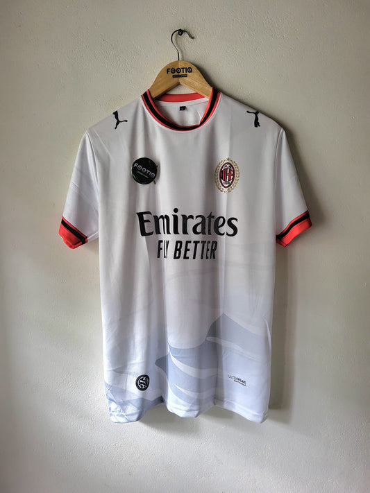 A white AC Milan soccer jersey with orange and black trim, hanging on a wooden hanger against a plain wall. The jersey features the Puma logo, AC Milan crest, Emirates sponsor, and a subtle blue and white wave pattern near the hem.