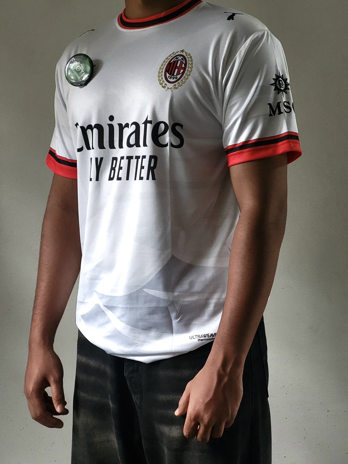 A person wearing an AC Milan soccer jersey with Emirates Fly Better branding, featuring a white body with red and black trim on the collar, cuffs, and sides.