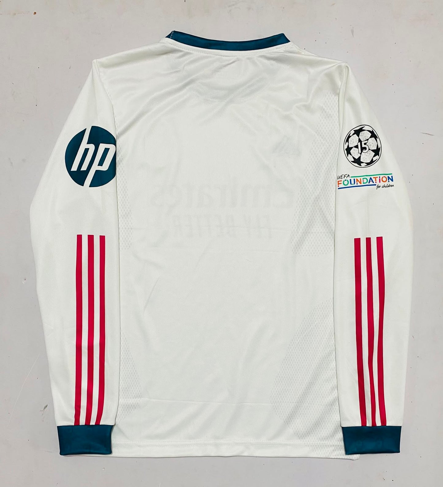 White long-sleeve soccer jersey laid flat on a light gray background, featuring dark teal collar and cuffs, red three-stripe detailing on the sleeves, an HP logo on the right sleeve, a Champions League patch on the left sleeve, and a Foundation by Adidas logo.