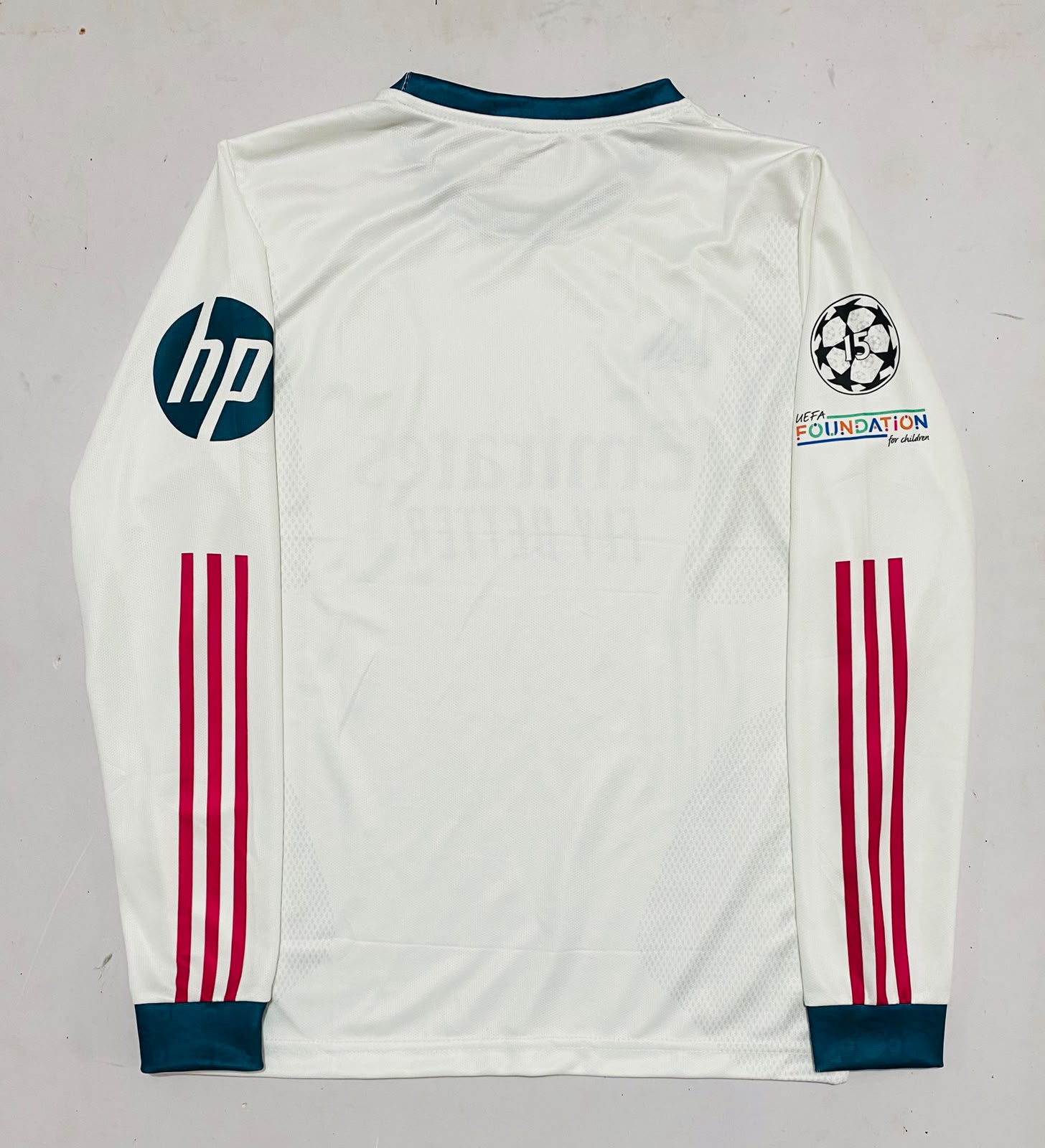 White long-sleeve soccer jersey laid flat on a light gray background, featuring dark teal collar and cuffs, red three-stripe detailing on the sleeves, an HP logo on the right sleeve, a Champions League patch on the left sleeve, and a Foundation by Adidas logo.