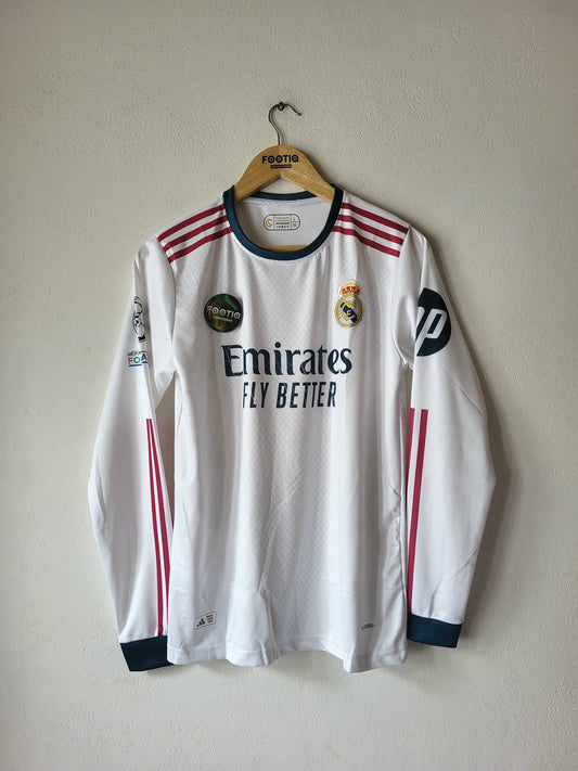 A white Real Madrid long-sleeve soccer jersey hanging on a wooden hanger against a plain beige wall. The jersey features navy blue trim, red stripes on the shoulders and sleeves, the Emirates Fly Better sponsor logo on the chest, and the Real Madrid club crest.
